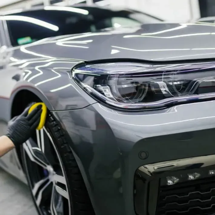Person wearing a black glove waxing a gray car's fender with a yellow applicator.