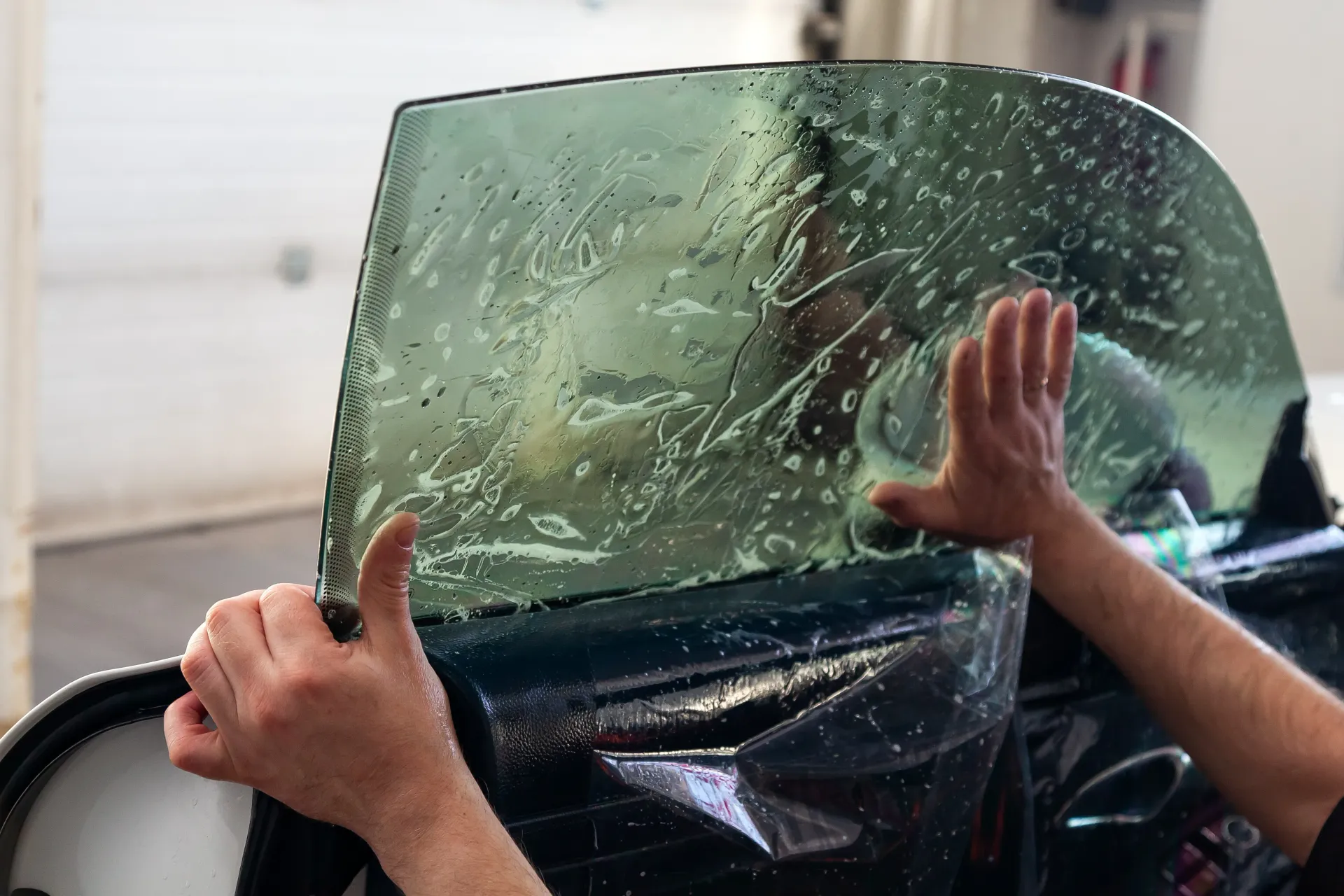 Person applying window tint to a car window; hands smoothing the film, with a light-colored garage setting.