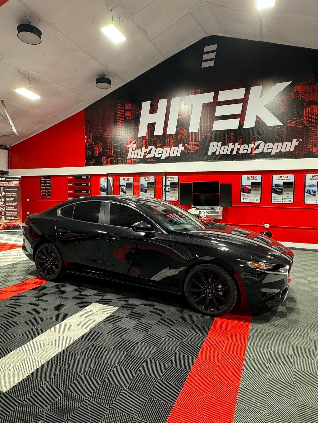 Black car inside a shop with a red and black graphic background, black wheels.