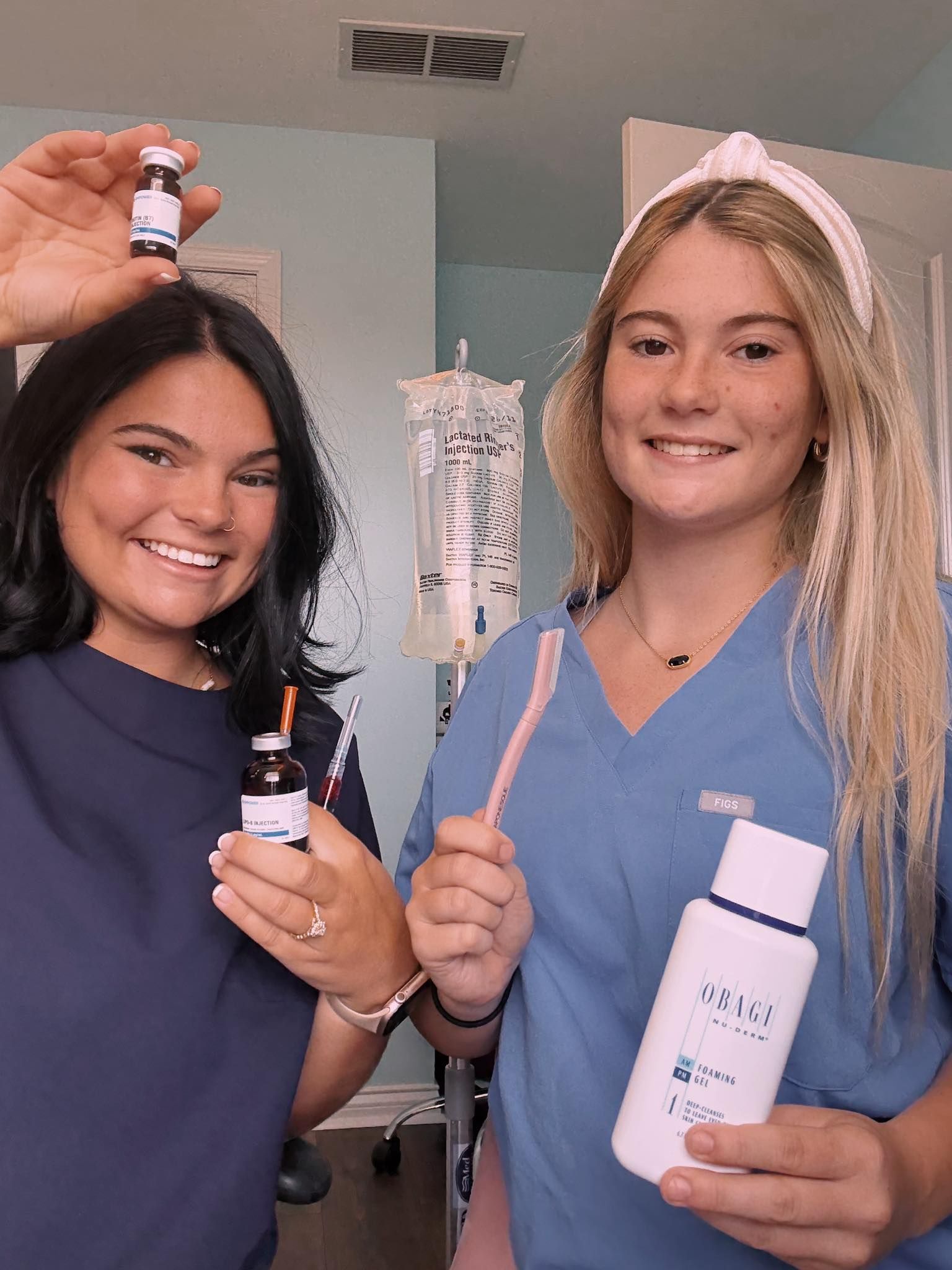 Two women in blue scrubs hold vials, toothbrush, and mouthwash, smiling in a room.
