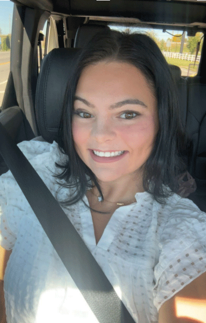Woman in white top smiling in a car, seatbelt on.