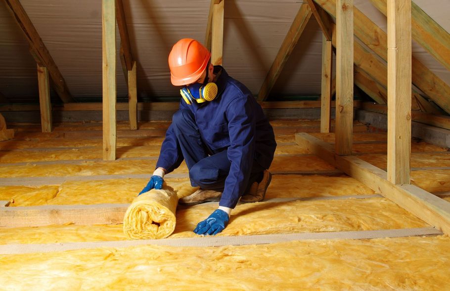 Worker laying yellow fiberglass insulation rolls in home attic.
