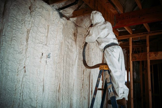 Worker on ladder applying expanding spray foam to interior wall studs.