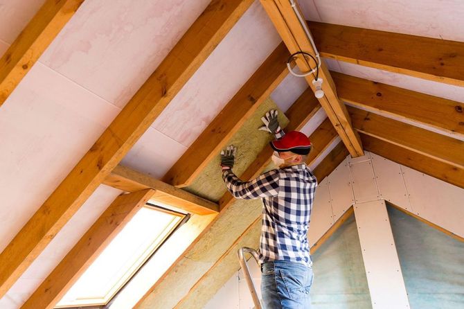Handyman installing fiberglass batt insulation between attic rafters.
