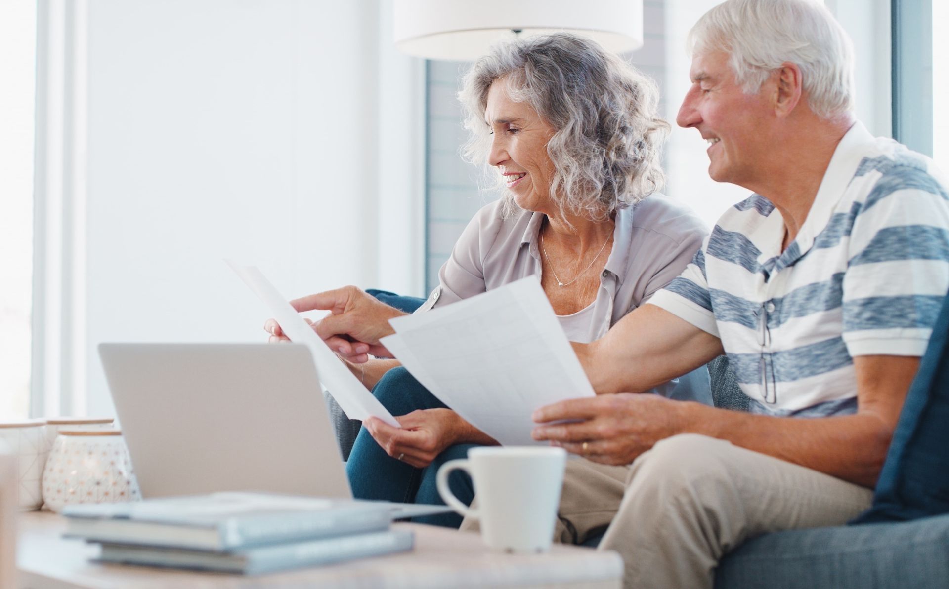 Senior couple reviewing papers together, pointing at document, smiling, sitting near laptop and coffee.