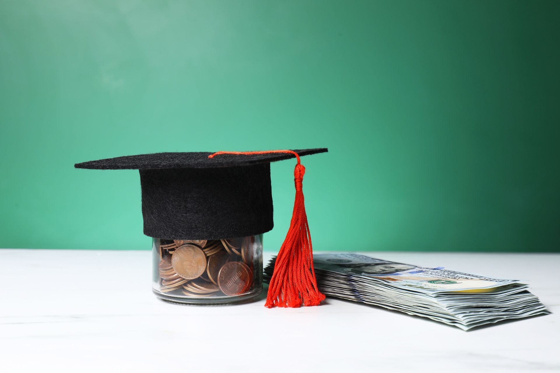 A graduation cap , a jar filled with coins and a stack of money on a table.