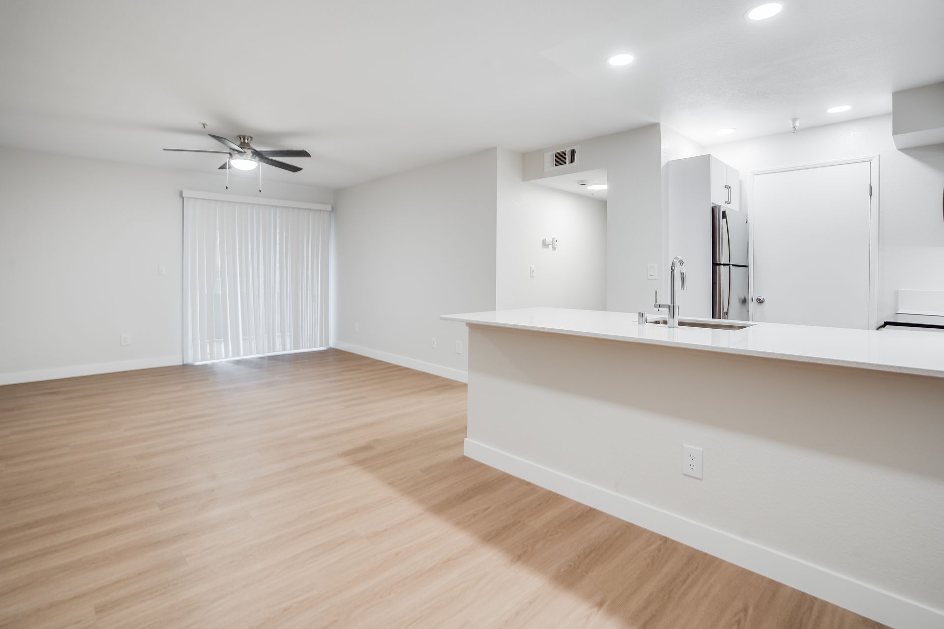 Empty, bright apartment interior with light wood floors, a white kitchen, and a sliding glass door.