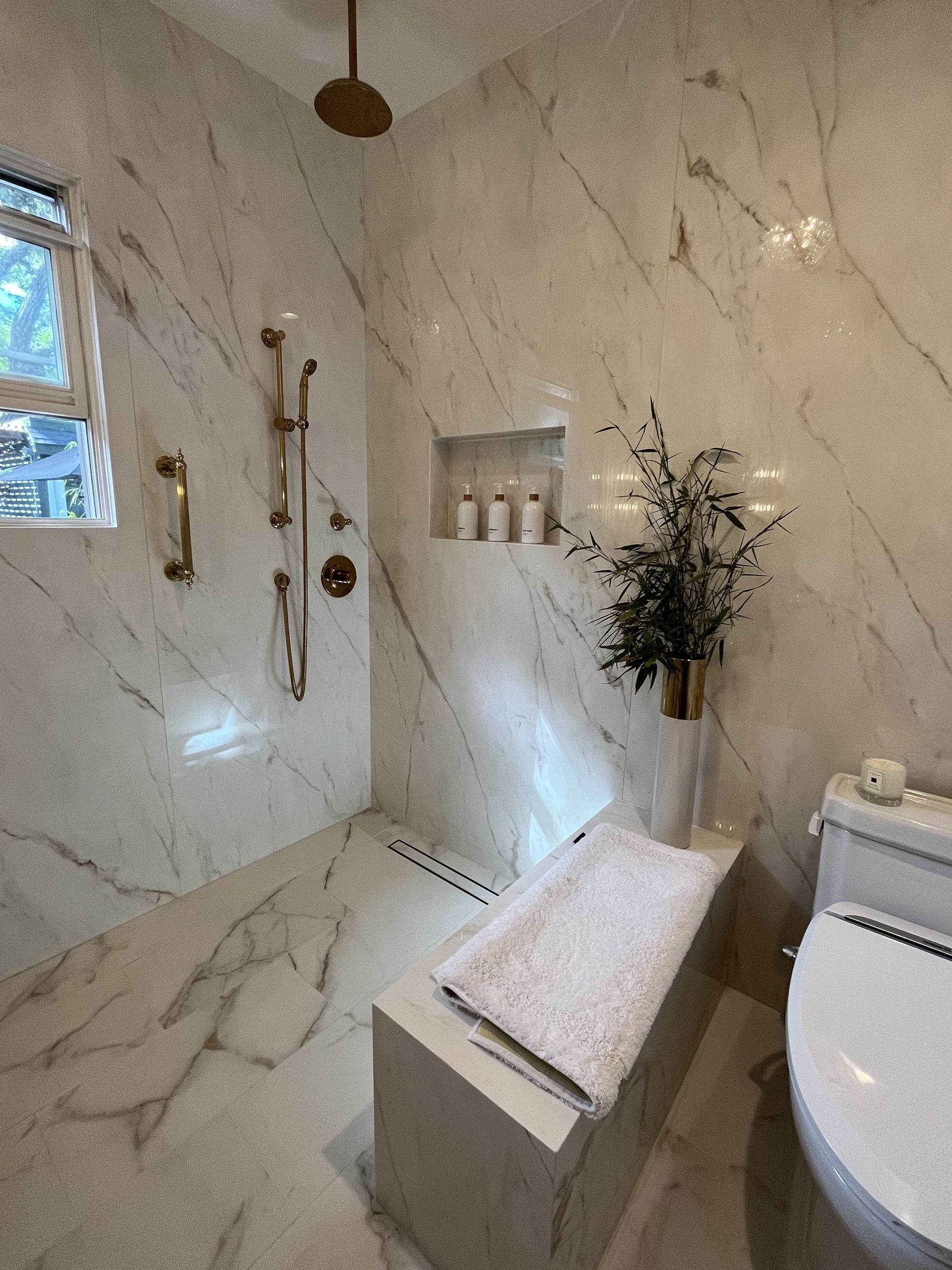 Luxurious white marble bathroom with gold fixtures. Shower, built-in bench, and toilet are visible.