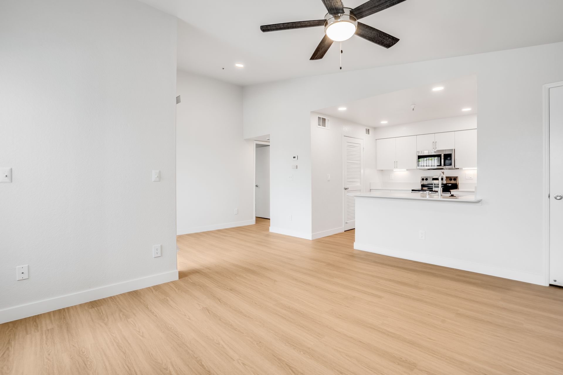 Empty living room with wood flooring, white walls, and a small open kitchen.
