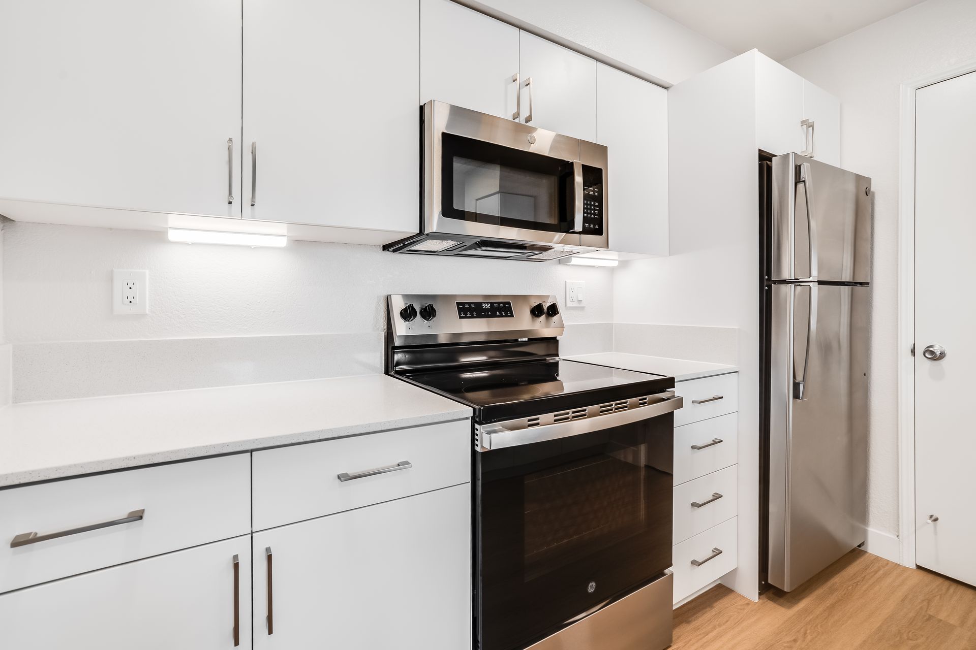 White kitchen with stainless steel appliances: microwave, stove, refrigerator. White cabinets, light wood floor.