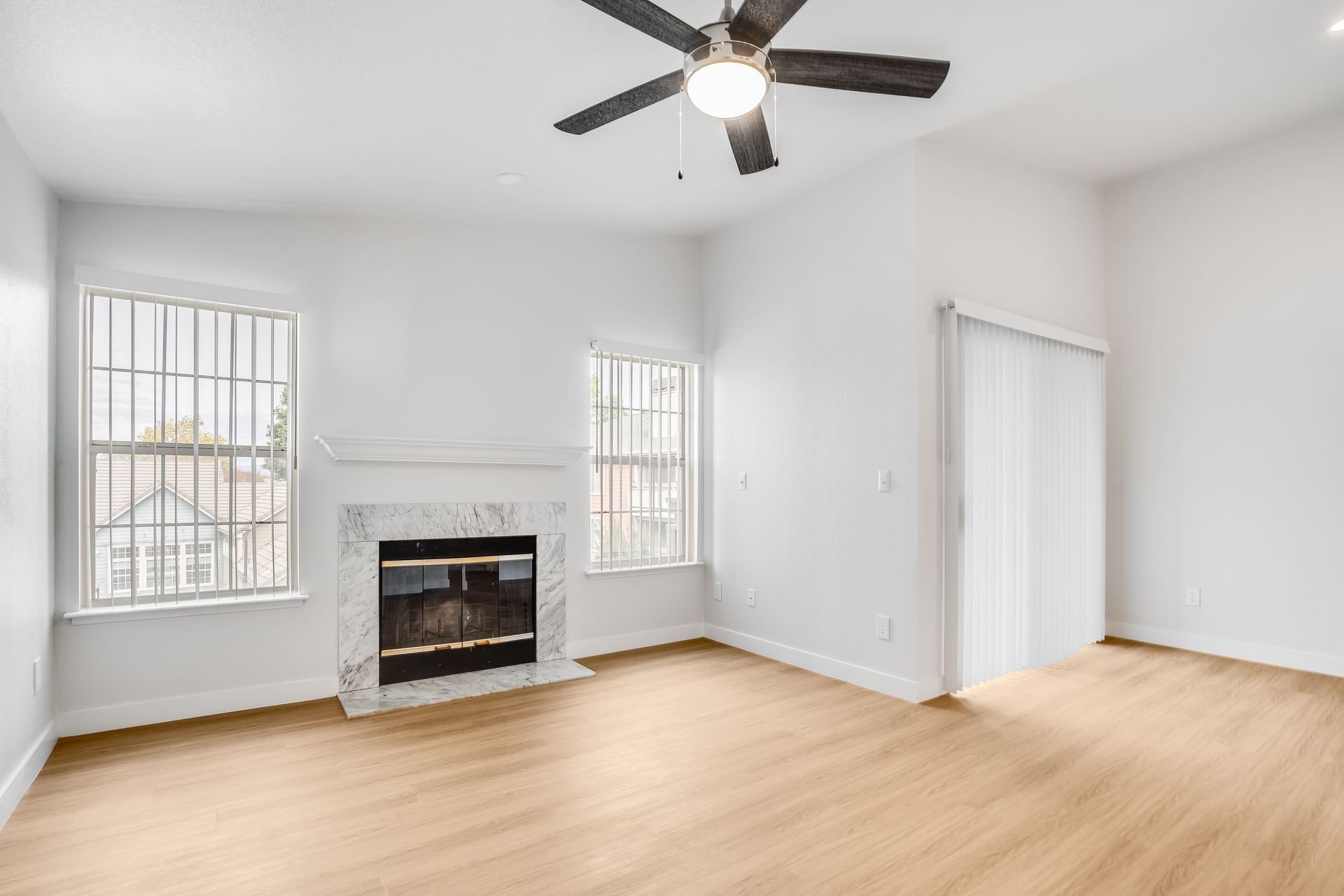 Empty living room with fireplace, windows, sliding door, ceiling fan, and light-colored wood flooring.