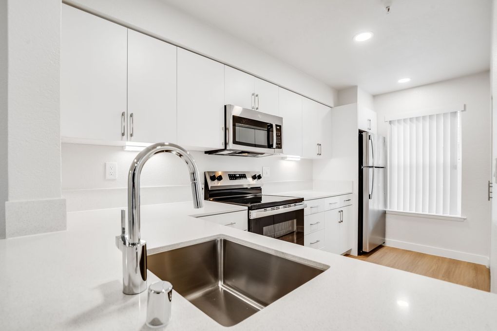 Modern white kitchen with stainless steel appliances, sink in the foreground, and a window.