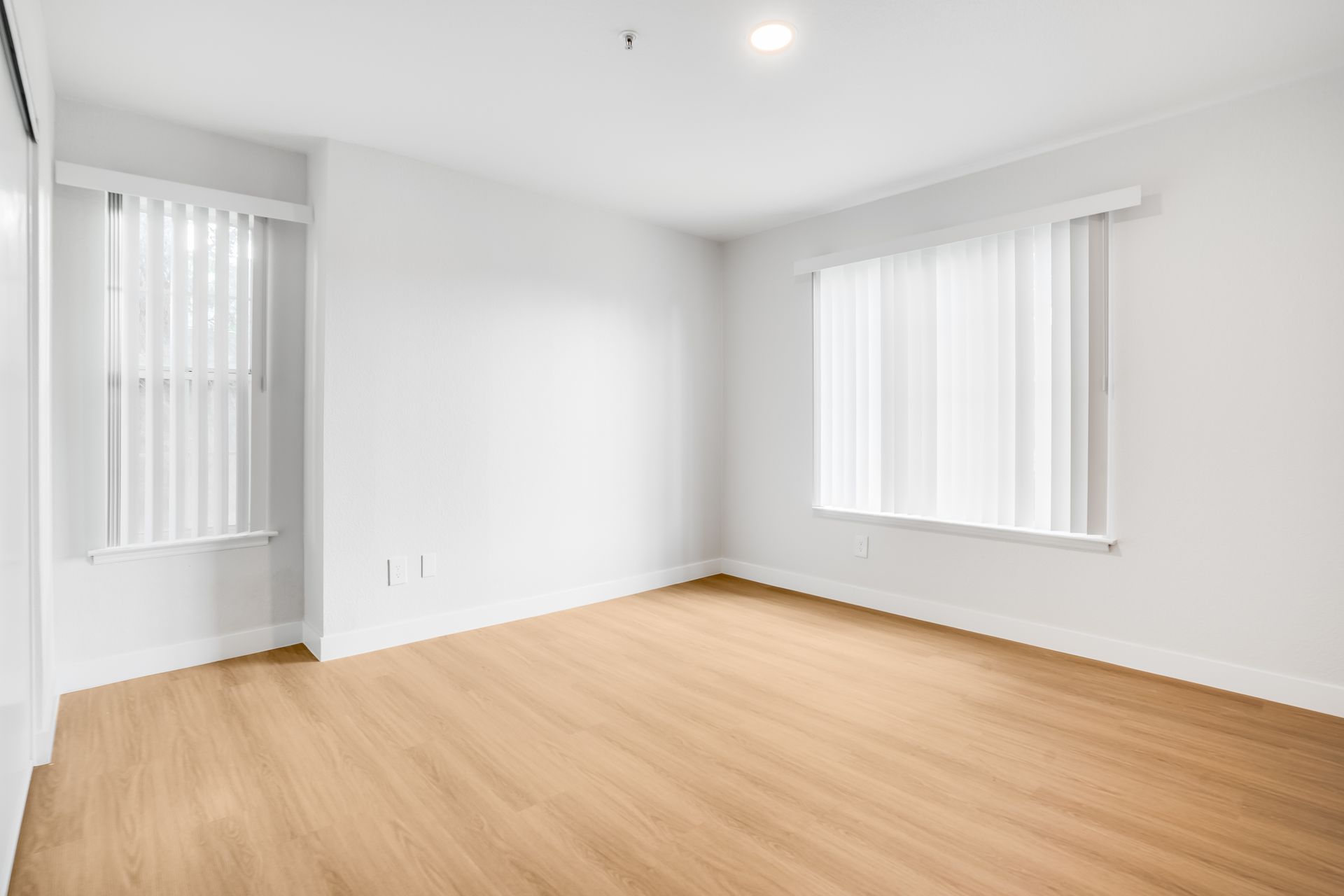 Empty room with hardwood floors, white walls, and two windows with blinds.