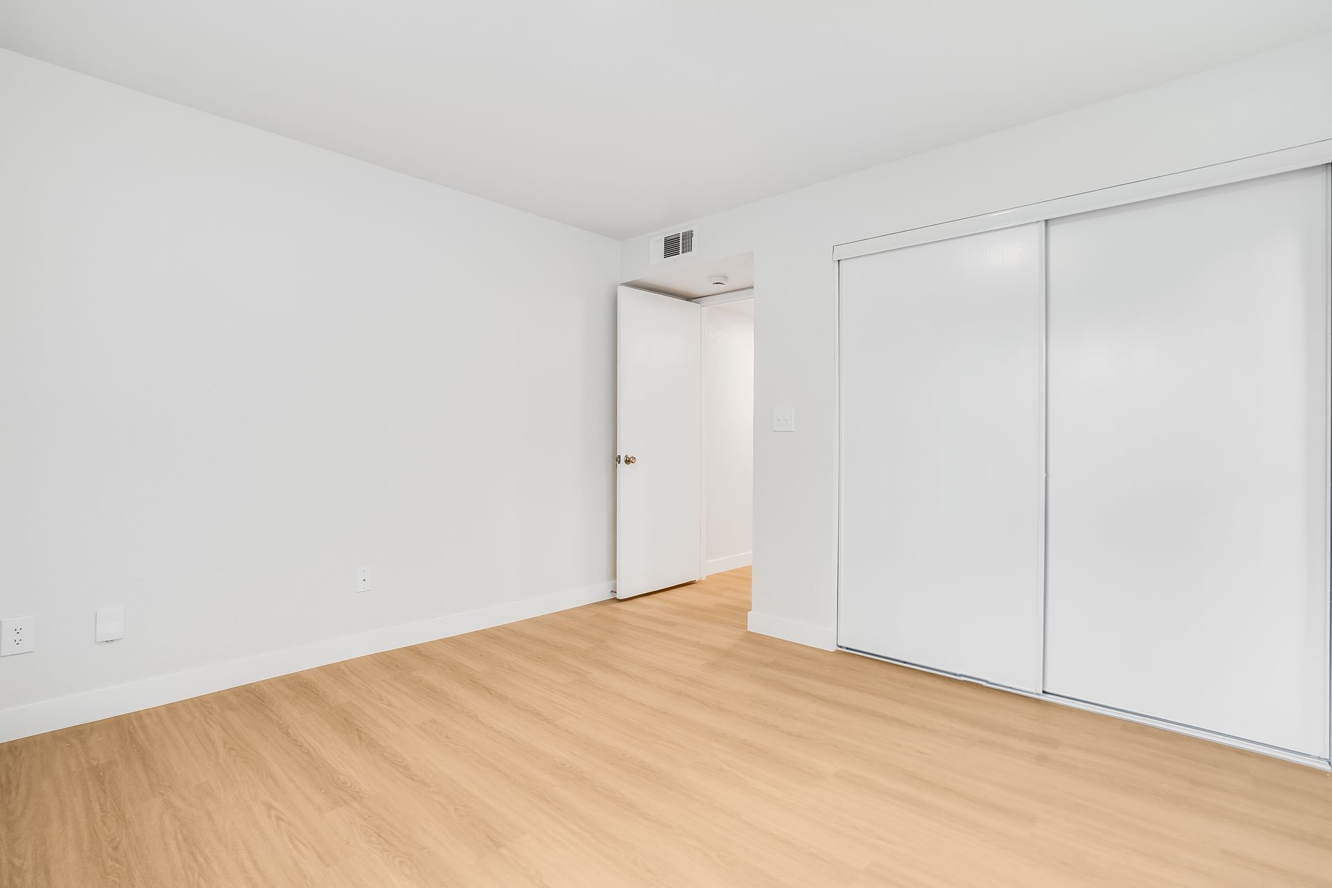 Empty room with white walls, light wood floor, sliding closet doors, and open doorway.
