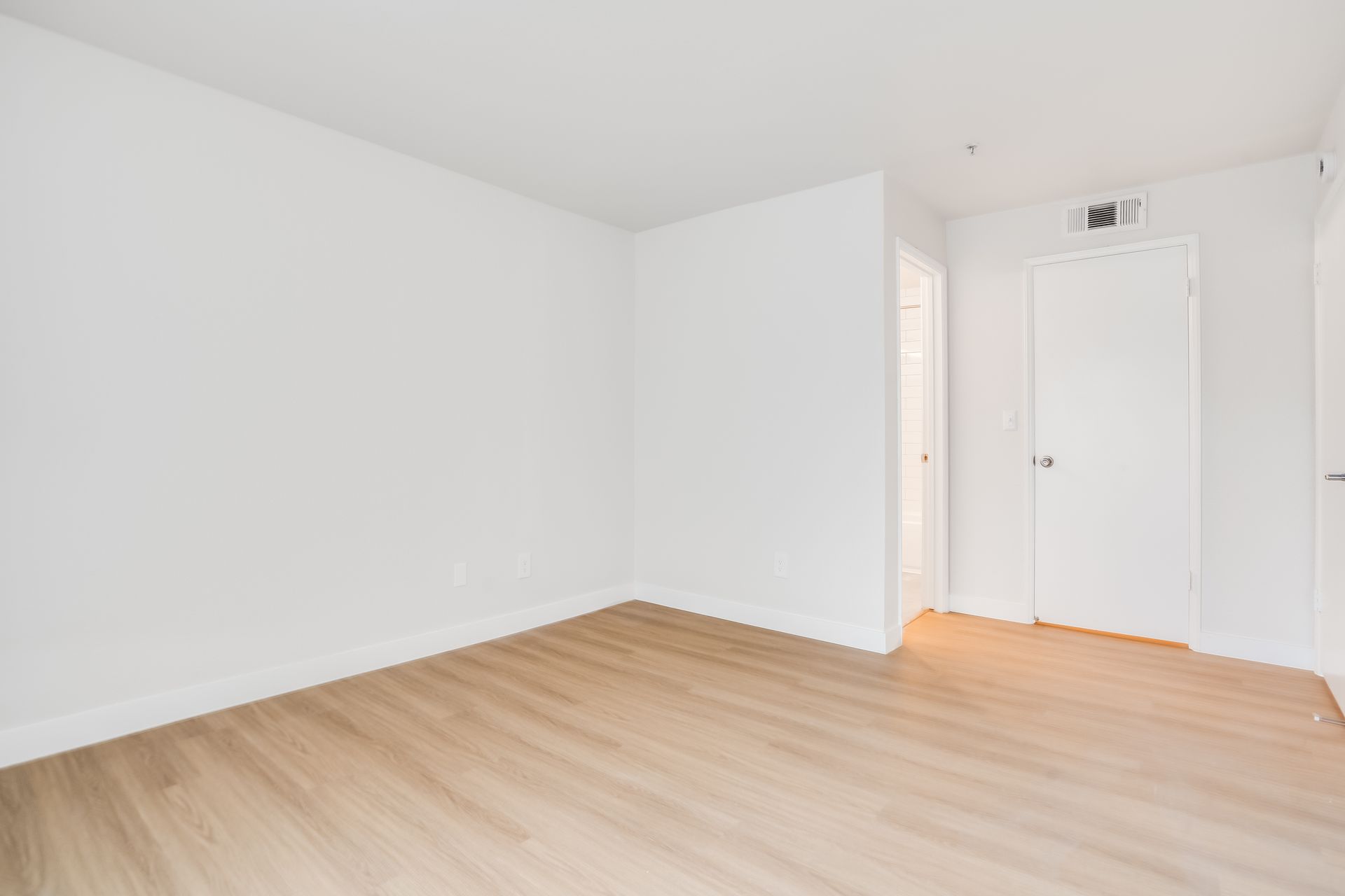 Empty room with white walls, light wood flooring, and a closed white door.