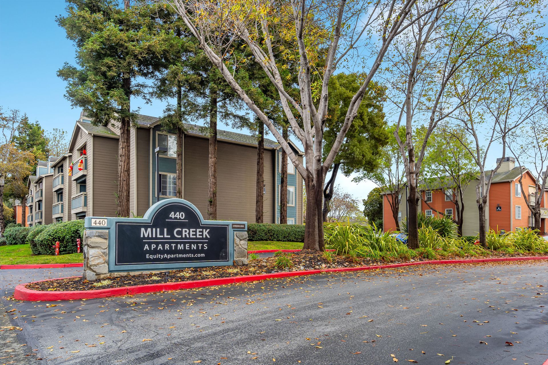 Sign for Mill Creek Apartments; multi-story building in background, trees and landscaping.