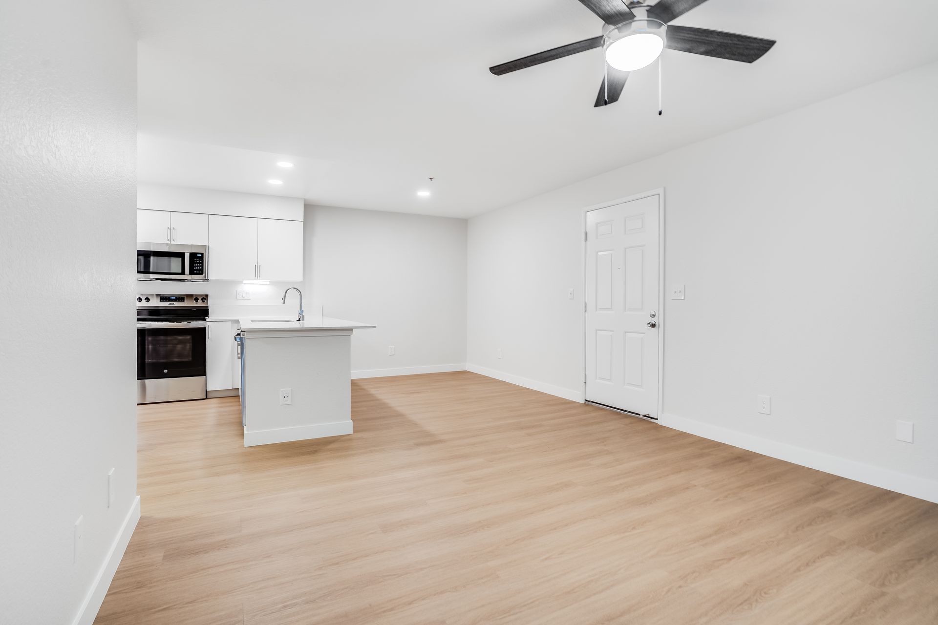 Bright, empty apartment interior with a kitchen, light wood floors, and a ceiling fan.