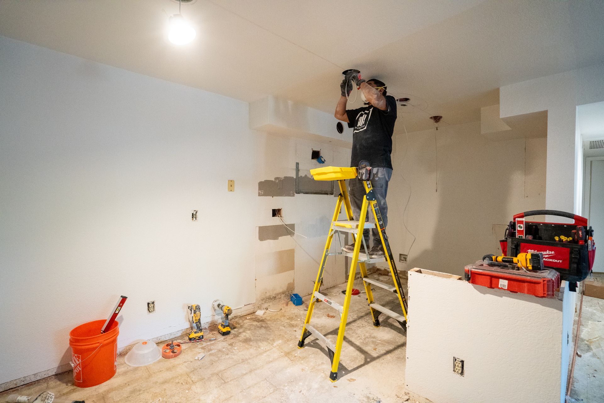 Person on a yellow ladder installing a light fixture in a room under construction.