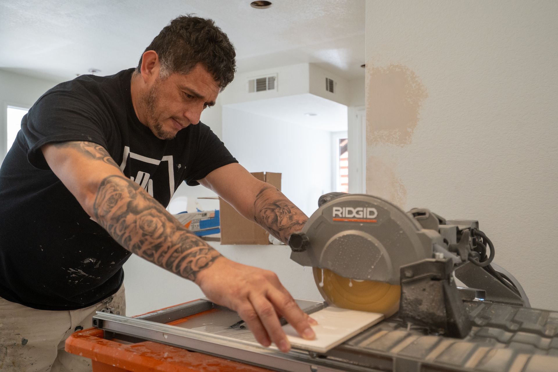 Man with tattoos cutting a tile with a power saw in a room under construction.