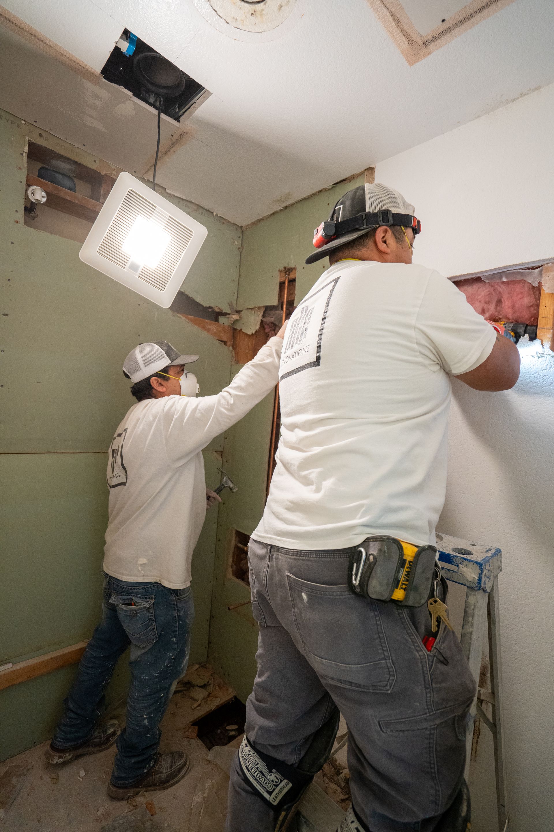 Two construction workers install a ceiling fixture in a room under construction.