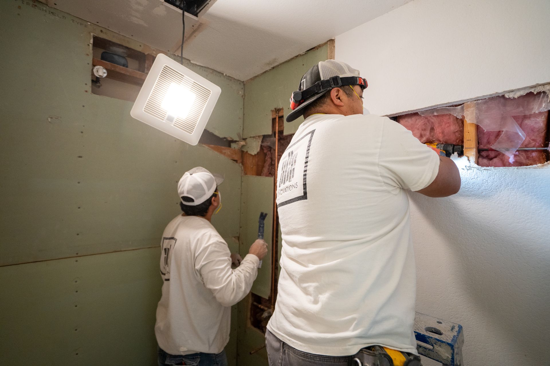 Two construction workers renovating a bathroom. One removes drywall, the other works on the wall.