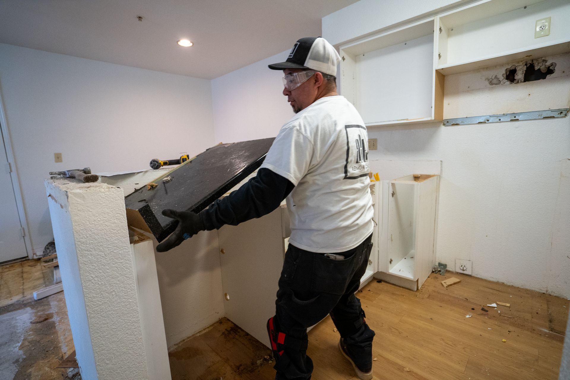 Man removing a countertop in a kitchen renovation; white shirt, black pants, gloves.