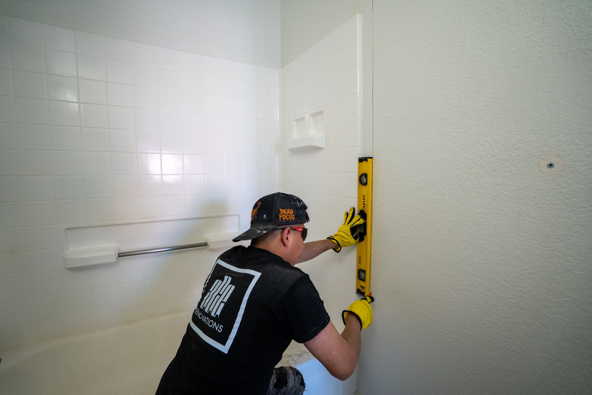 A person in a black shirt uses a level to check a shower wall during installation. Yellow gloves. White shower.
