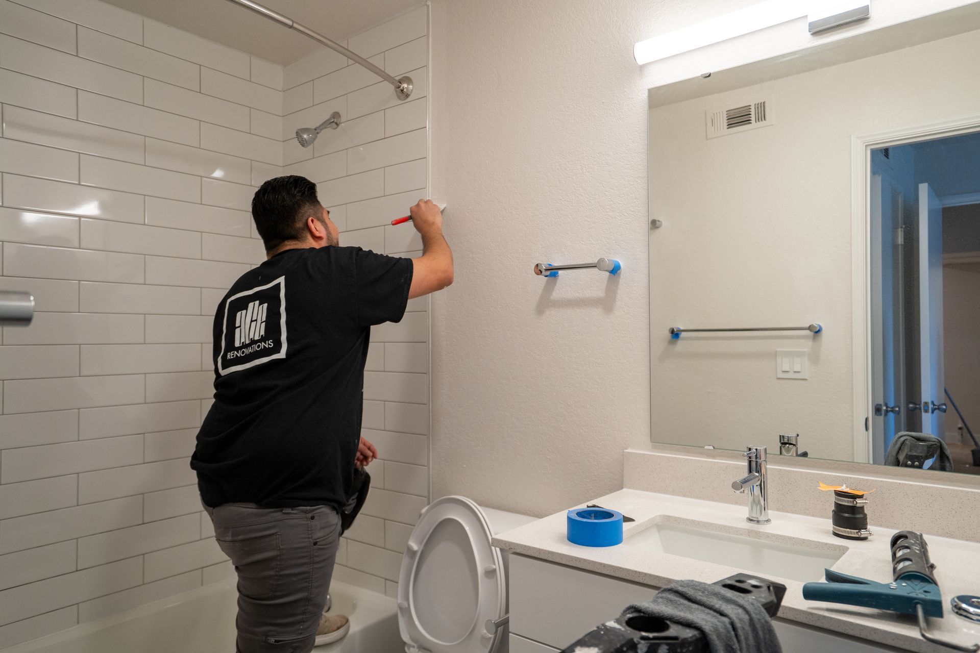 A person paints a bathroom wall, near a shower, sink, and toilet.