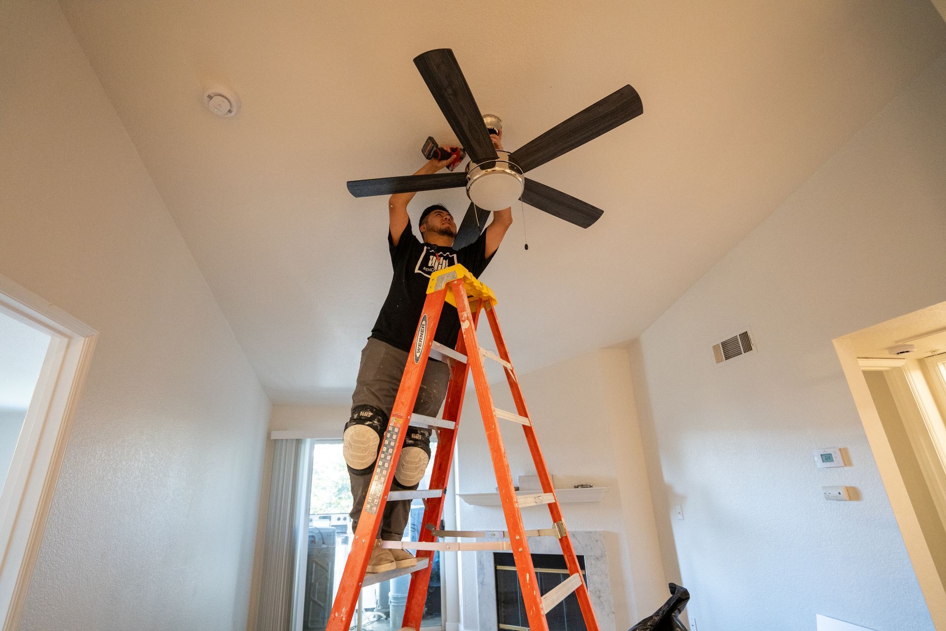 Person installing ceiling fan on a ladder in a brightly lit room.