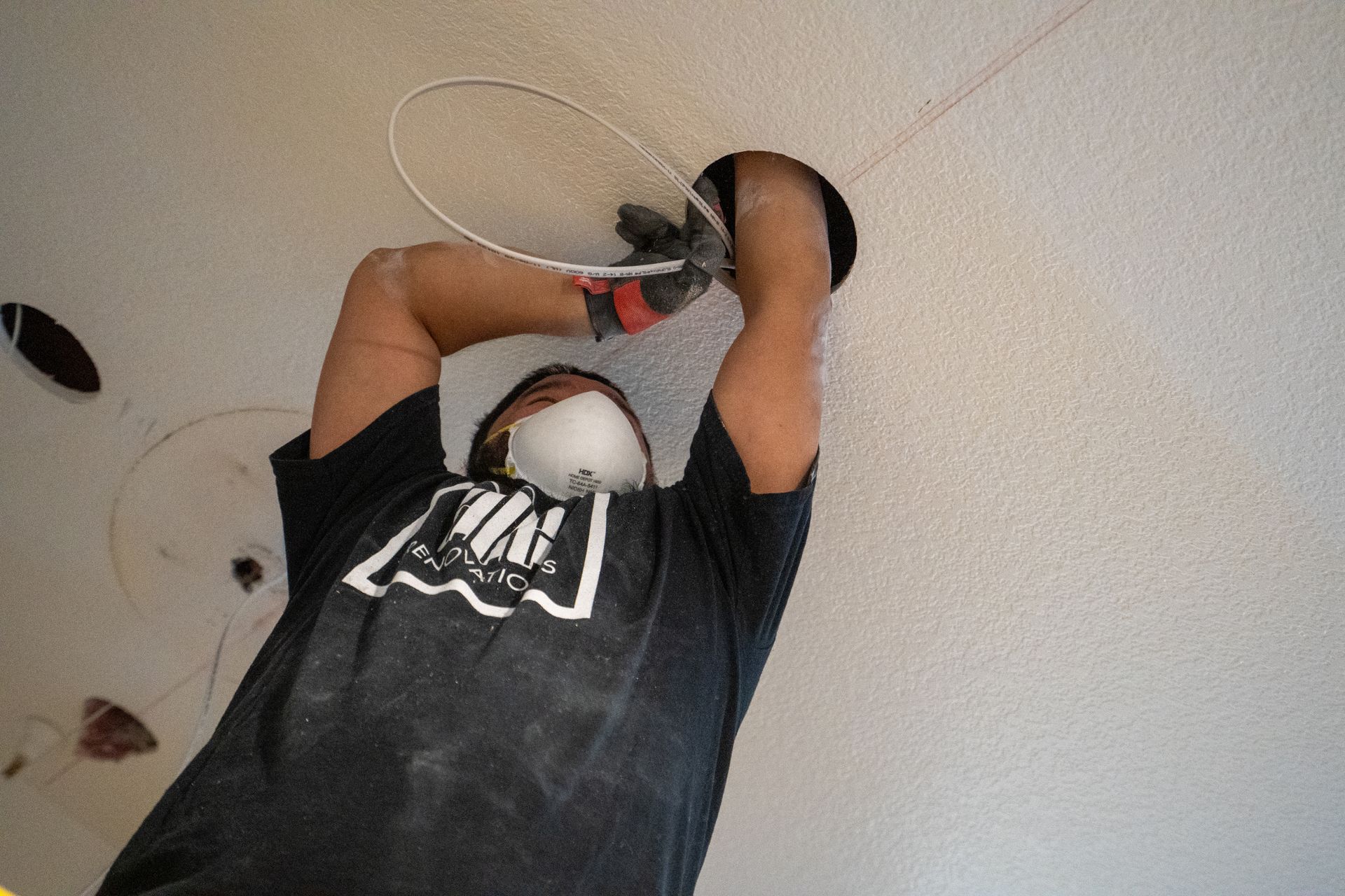 Person installing a fixture in a white ceiling, wearing a mask and gloves.