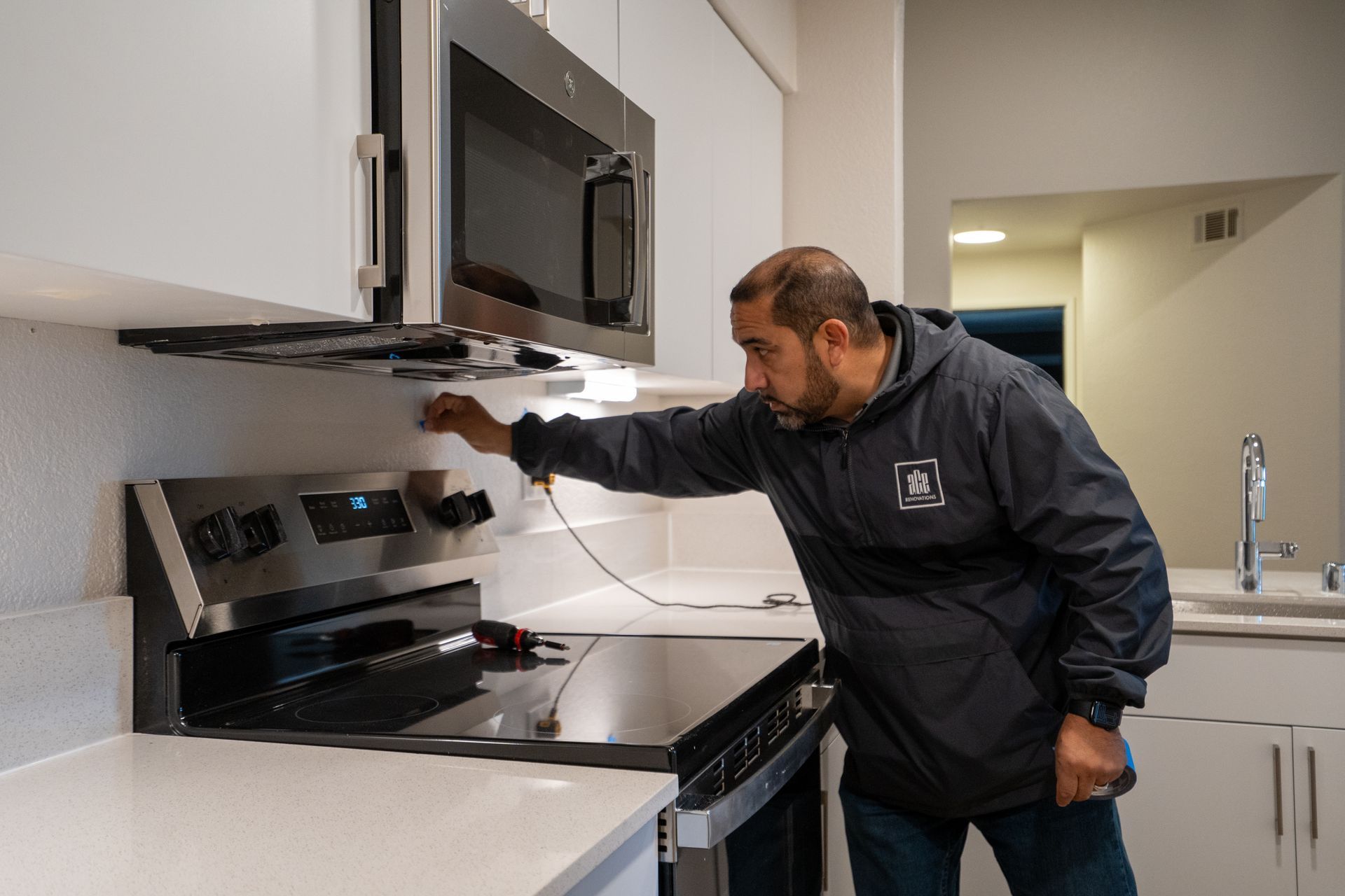 Man in kitchen inspecting stovetop and microwave.