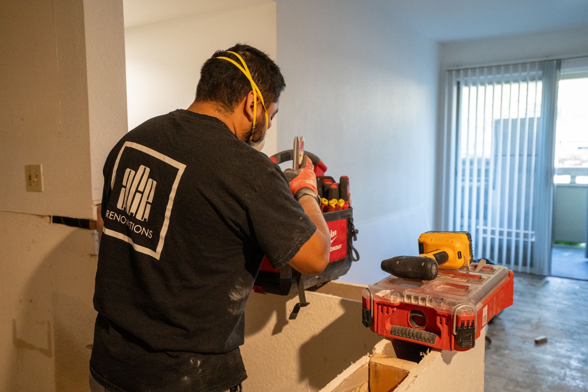 Man wearing safety glasses and a mask using a tool to work on a wall, with tools nearby in a room.