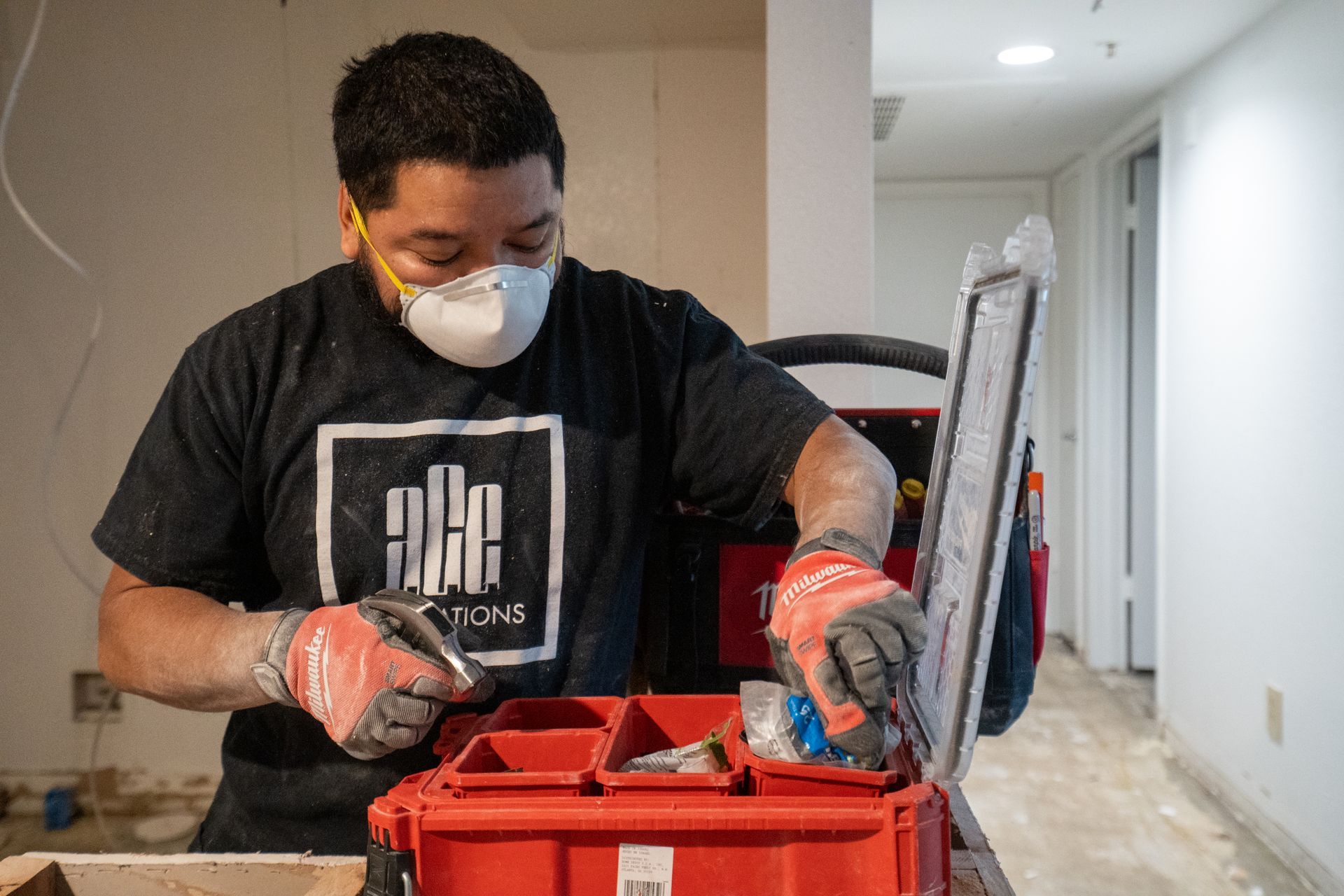 Construction worker in mask and gloves, looking in a red toolbox in a room under renovation.