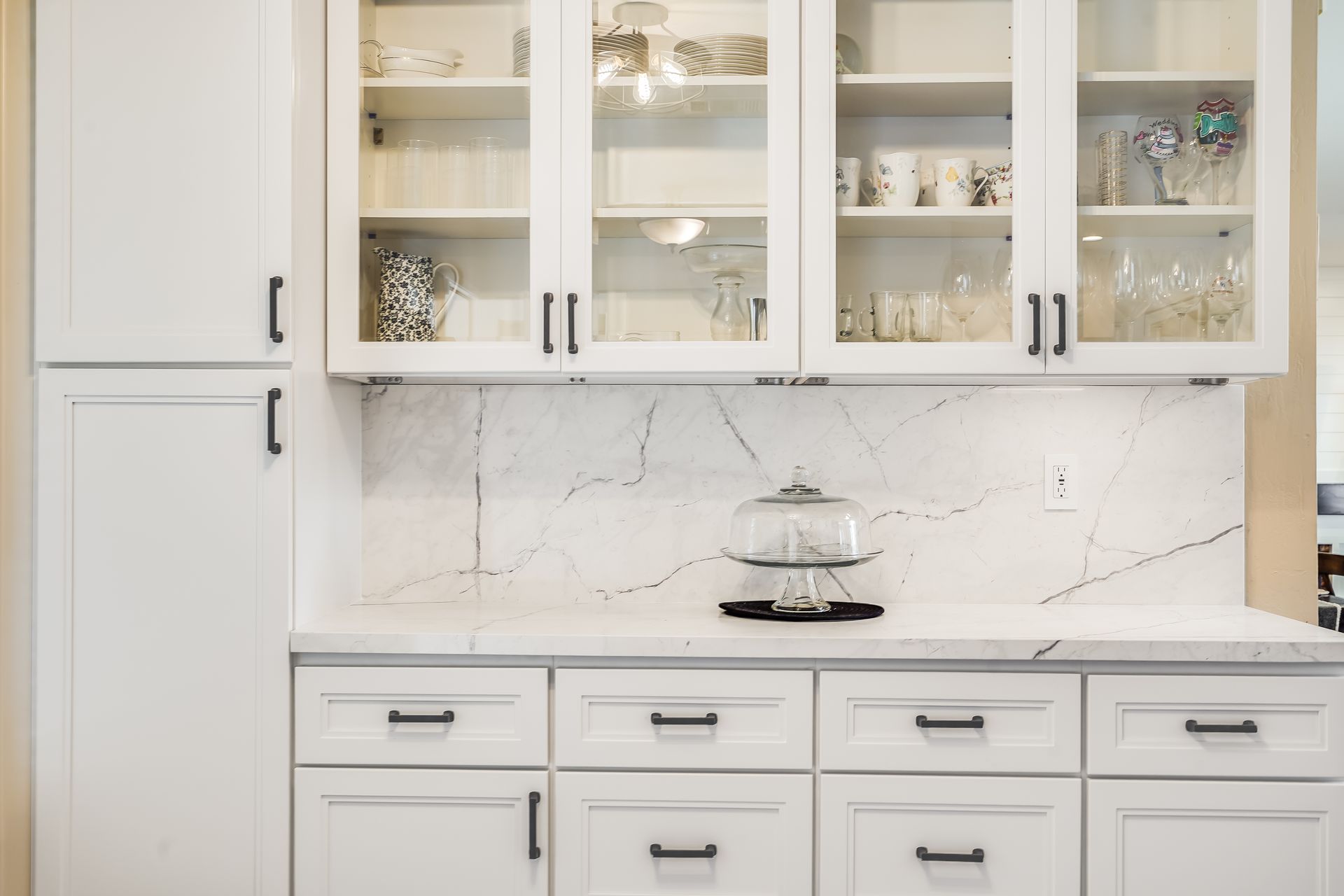 White kitchen cabinets with glass-front uppers, black hardware, and marble backsplash.