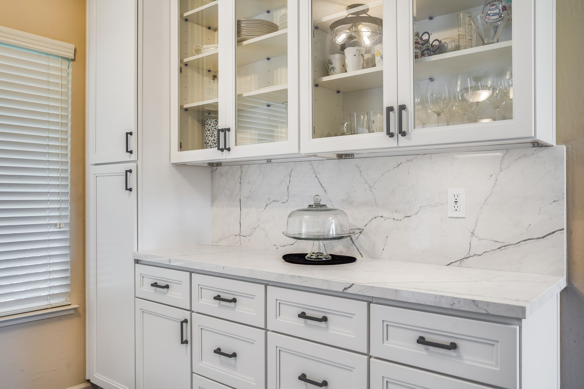 White kitchen cabinets with glass-front uppers, countertop, and a decorative cake stand.