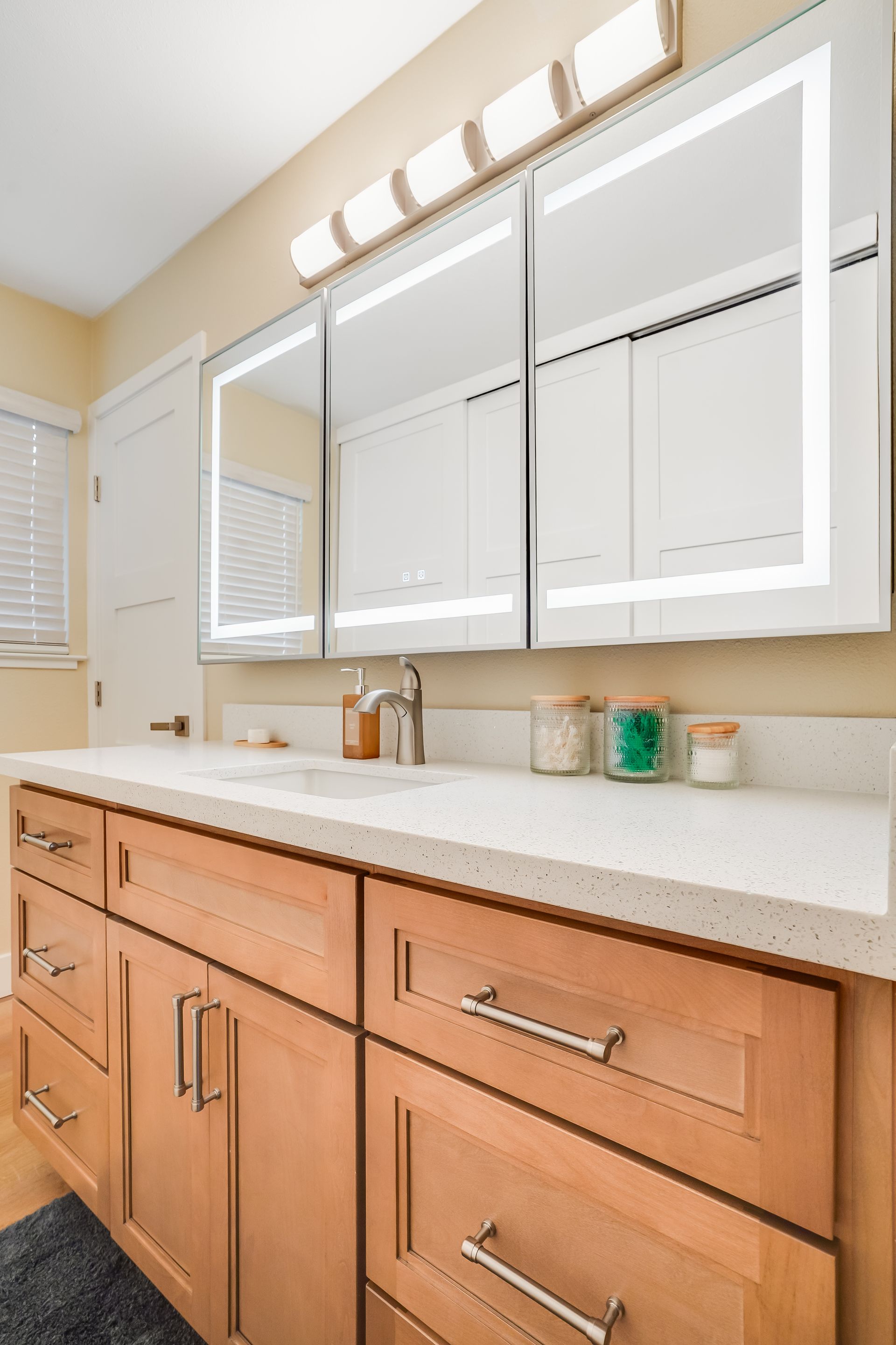Bathroom vanity with light-up mirrors, light-brown cabinets, and a white countertop.