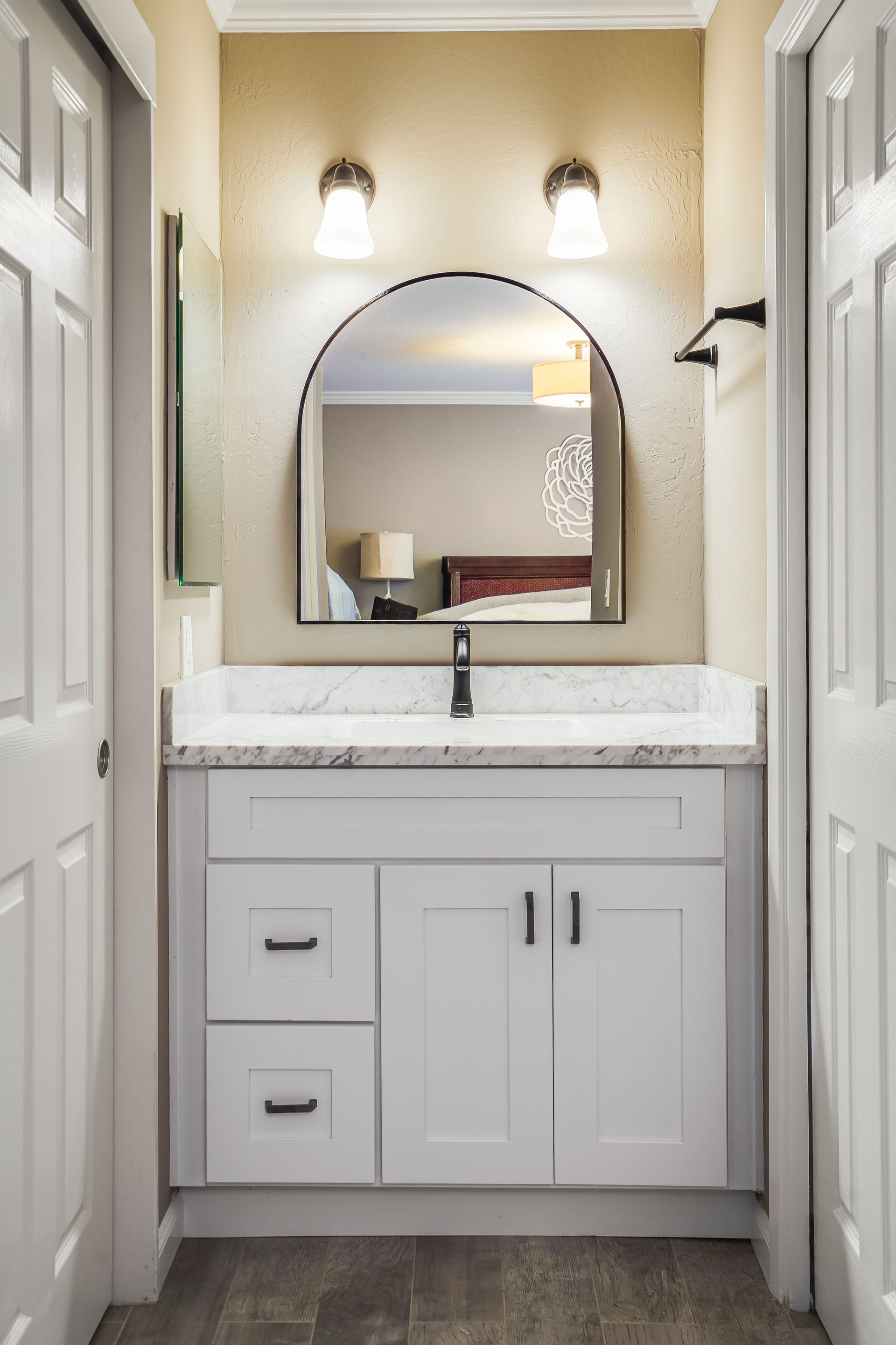 White bathroom vanity with arched mirror, two sconces, and closed white doors on either side.