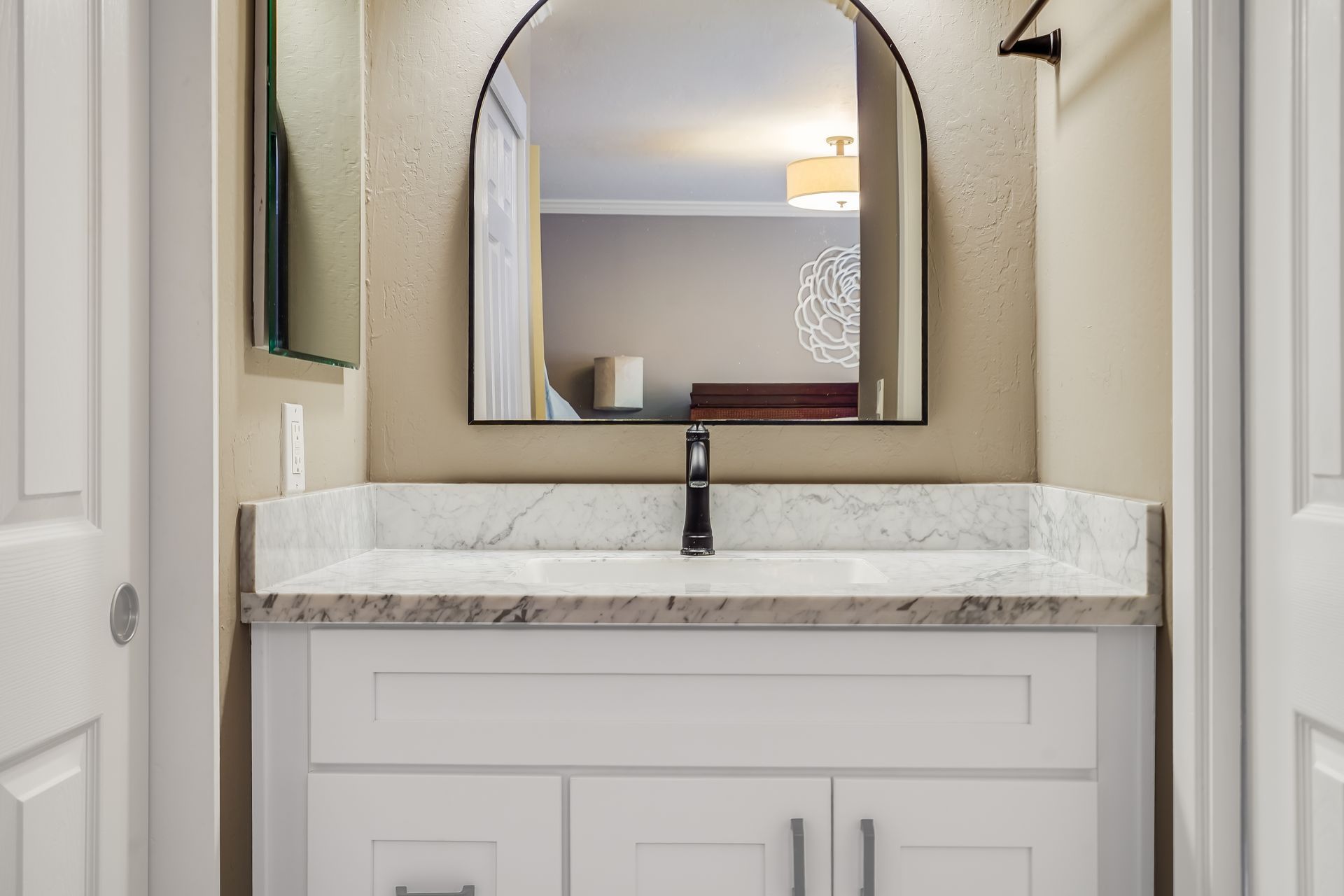 White bathroom vanity with marble countertop, black faucet, and arched mirror.