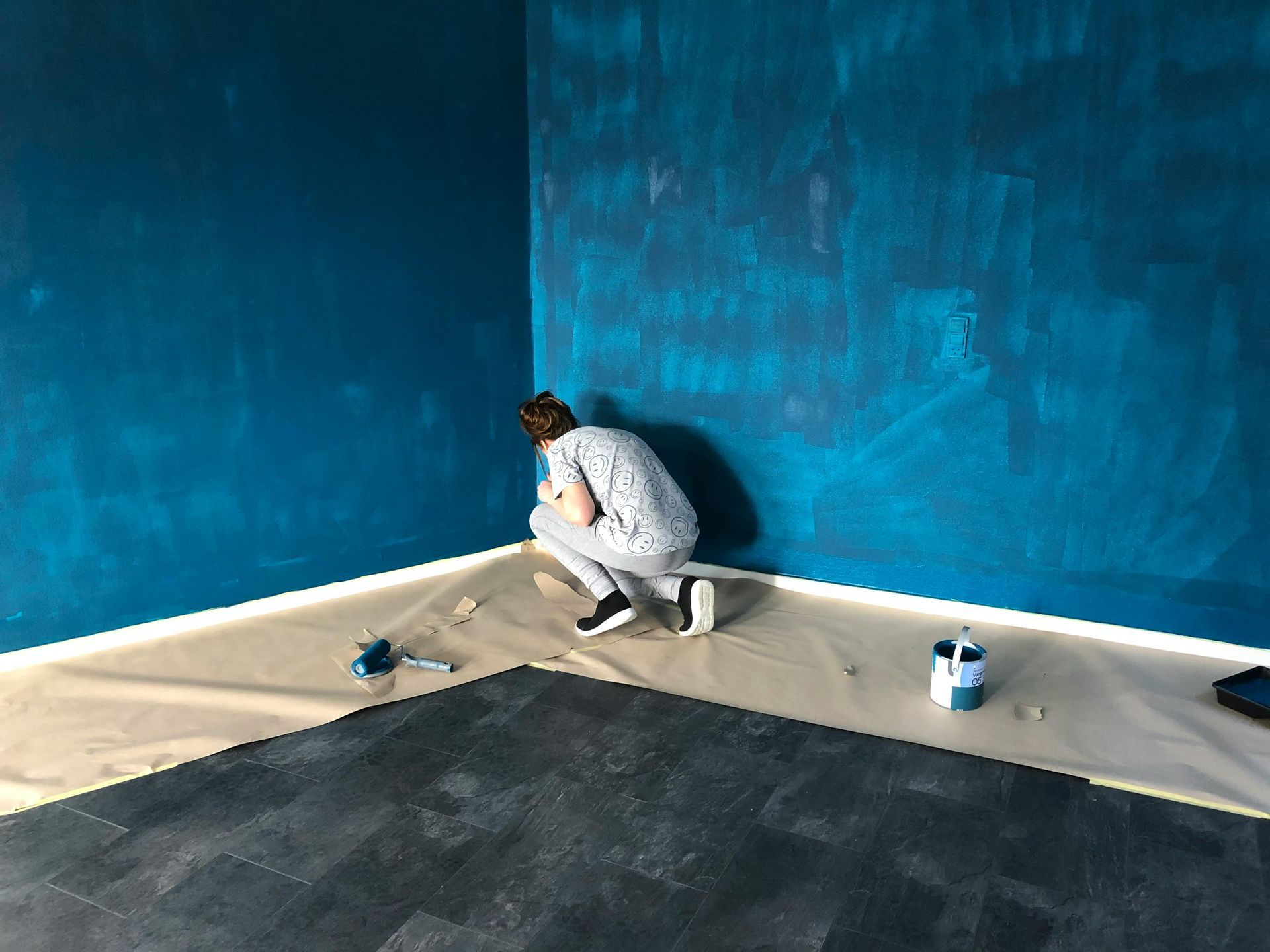 Person painting a wall in dark teal; painting supplies on floor.