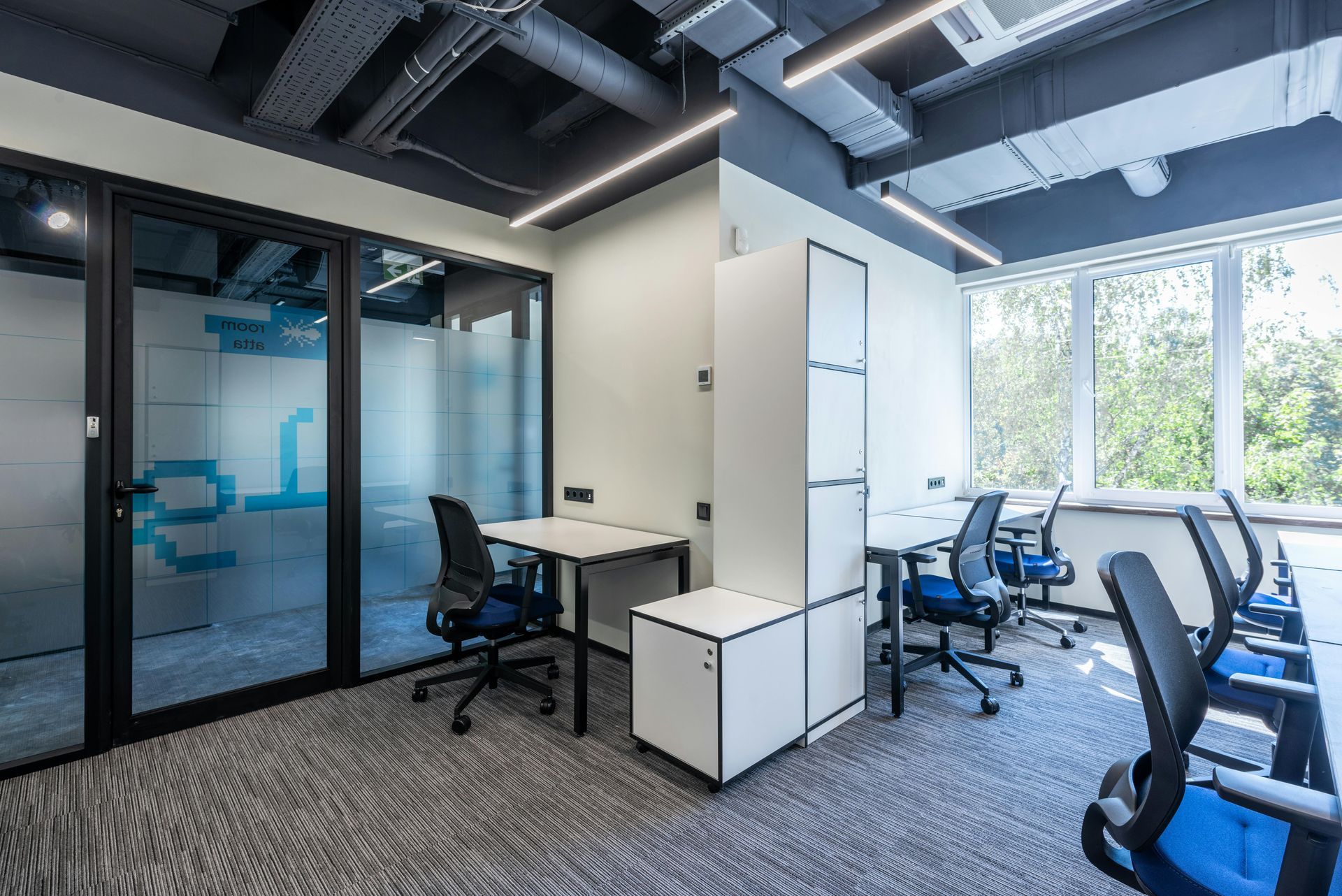 Modern office space with desks, chairs, glass-walled room, and large windows with natural light.