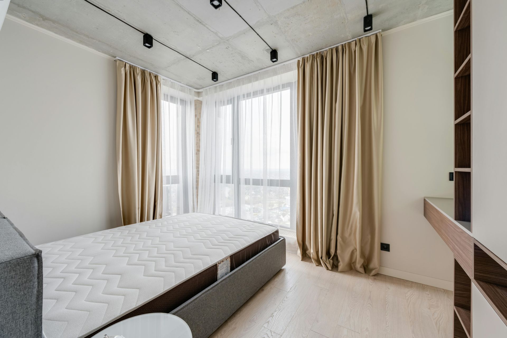 Bedroom with tan curtains, a single bed, and a built-in shelf unit. Light wood floors and a concrete ceiling.