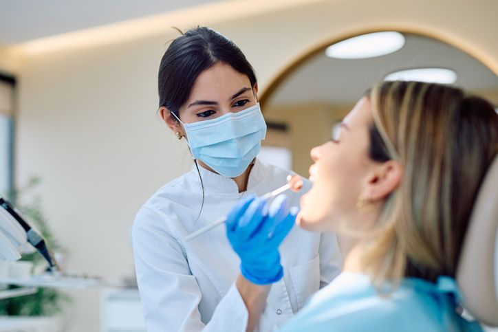 Dentist examining a patient’s mouth in a bright clinic, both wearing masks and gloves.
