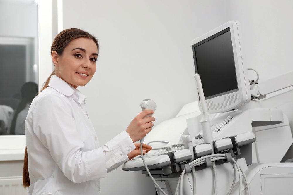 Sonographer holding an ultrasound probe beside a patient chair and monitor in a clean clinic room
