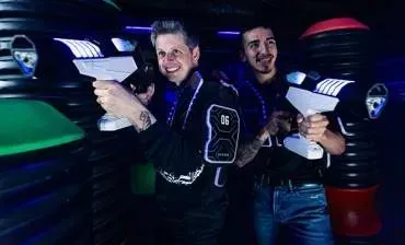 Two people smiling at a neon-lit arcade laser tag scene, aiming white laser tag guns