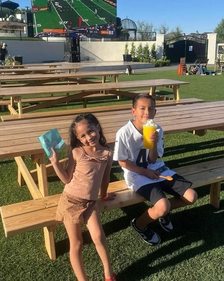Two children sitting on a picnic bench, smiling and holding colorful drinks at an outdoor stadium.