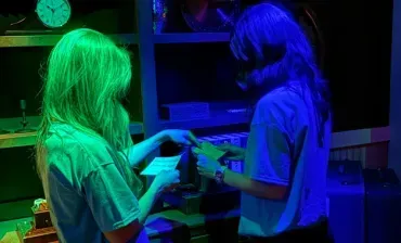 Two people in neon green and blue light exchange an object in a dark room.