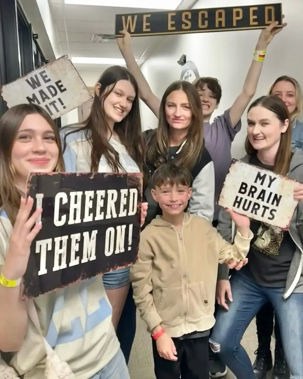 Group of smiling people holding “WE ESCAPED,” “WE MADE IT,” and “MY BRAIN HURTS” signs at a celebration.