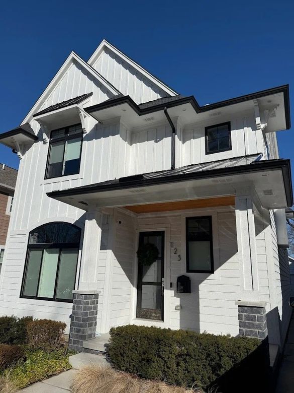 White two-story house with black trim, front porch, and landscaped shrubs under a clear blue sky