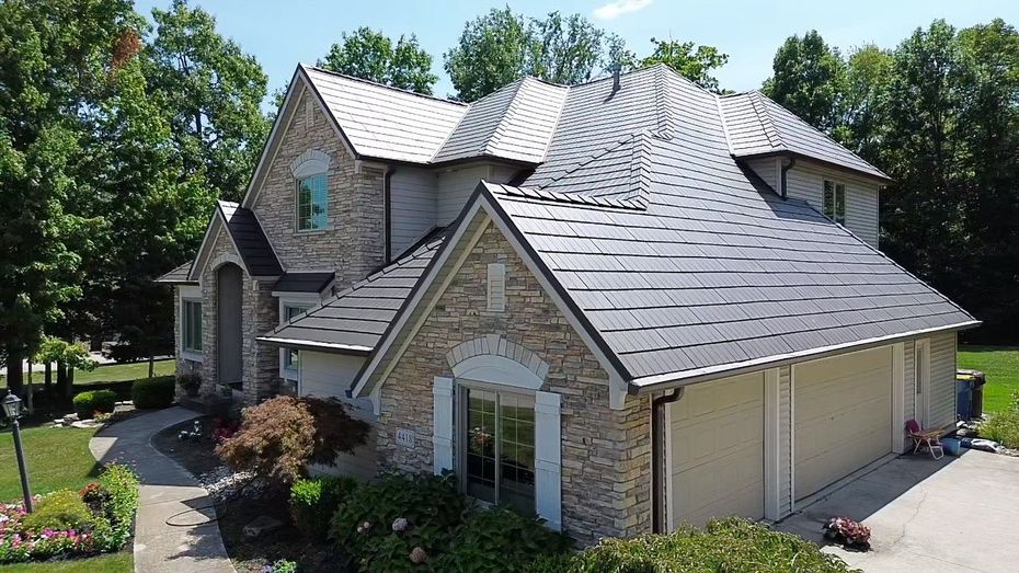 Stone suburban house with gray shingled roof, arched entry, and beige garage surrounded by trees