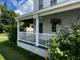 White porch with railings on a light gray house, hanging flower baskets, and a green lawn under a blue sky